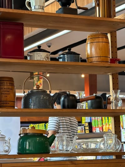 This image depicts an indoor commercial setting in Benoa, Indonesia, specifically a retail display featuring multiple wooden shelves laden with various coffee and tea accessories.

**Environment:** The background reveals a retail space with illuminated shelving displaying packaged goods, likely food or beverage products. Overhead, ceiling lights and a small, black security camera are visible. A partial round sign indicating "10%" is also discernible. The structure consists of sturdy, light-colored wooden shelves, approximately four tiers deep.

**Subjects/Objects:**
*   **Top Shelf:** Contains a white and silver Moka pot, a black manual coffee grinder with a wooden handle, and a tall, brass-colored cylindrical container.
*   **Second Shelf:** Features a dark red, rectangular metal tin; a small white ceramic pot with a black lid, adorned with "taste enjoy relax" text; a similar dark blue ceramic pot; a small wooden barrel with black bands; and a small red cylindrical object.
*   **Third Shelf:** Displays a wooden cylindrical container with text identifying "BLUE MOUNTAIN COFFEE PRODUCT OF JAMAICA"; a dark grey, speckled metal teapot with a wooden handle; two black metal gooseneck pour-over kettles, one with a wooden handle; and two clear glass decanters of differing sizes.
*   **Bottom Shelf:** Holds a green enamel teapot with a wooden lid, and various clear glass teapots, infusers, cups, and saucers.
*   **People:** One individual is partially visible from the back, positioned behind the shelves and to the right of the green teapot on the bottom shelf. The person is wearing a short-sleeved shirt with horizontal white and black stripes.

**Actions/Interactions:** No active interactions or specific actions are discernible; the scene primarily presents a static display of merchandise with a person passively present in the background.
