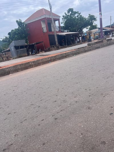 The image captures a street scene in Kona Garu, Nigeria, with a focus on a red building and the adjacent road. The perspective is from the side of the road, looking towards the building and across the asphalt.
**Subjects:**
The scene includes a two-story red building with white trim, featuring balconies with black railings on the upper floor. In front of the building, there are several smaller structures with corrugated metal roofs, some appearing to be stalls or extensions. Trees with dense green foliage are visible behind and around the buildings. The road is paved with asphalt and shows some signs of wear and tear, with scattered dark spots. A few people are visible near the structures, some sitting.
**Activity or Event:**
The presence of people suggests some form of daily activity, possibly commerce or resting. It does not appear to be a major event.
**Notable Details:**
The time of day seems to be daytime, given the lighting. The sky is overcast, indicating cloudy weather. There are power lines visible, suggesting an urban or semi-urban environment. The overall impression is of a somewhat modest and possibly developing area.
**Visible Text:**
Text is visible on a purple pole in the background, but it is too blurry to be legible. There are also some yellow signs on a building in the background with some writing, but it is also not clear enough to read.
