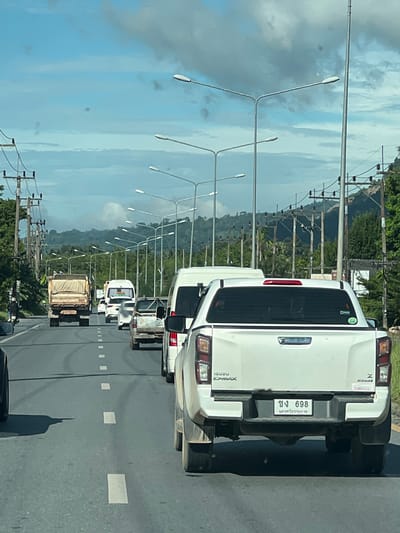 The image depicts a traffic scene on a multi-lane road in Ban Put Hong, Thailand. In the foreground, a white pickup truck with a Thai license plate "ขง 698" is visible, facing away from the camera. The truck has "ISUZU D-MAX" branding on its tailgate. To the left of this truck, another white pickup truck's side mirror is visible. Further ahead, several other vehicles are in traffic, including a white van, a truck with a tan cover, and smaller cars. The road is paved and marked with white dashed lines. On the sides of the road, numerous tall streetlights are installed. Utility poles with electrical wires run along the roadside, disappearing into the distance. The background features a lush green landscape with rolling hills and mountains under a partly cloudy blue sky. The overall scene suggests a typical day with moderate traffic flow.