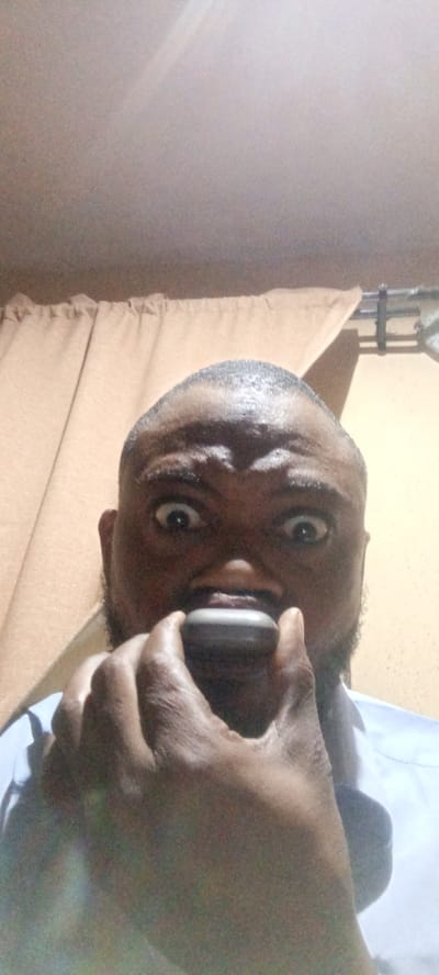 A close-up, low-angle portrait captures a dark-skinned man with a short buzz cut and a beard, looking directly at the viewer with wide, exaggerated eyes that convey surprise or a playful expression. He is wearing a light-colored, possibly blue or white, collared shirt, visible on his shoulders and chest.

The man is holding a small, dark grey or black, oval-shaped object, likely a jaw exerciser or a similar oral device, with his fingers positioned against his mouth. The activity appears to be the use of this device, possibly for exercise or as a prop for his expressive pose.

The setting is indoors, with a light brown or beige curtain visible in the background, secured by a dark curtain rod. The lighting is artificial, suggesting an indoor environment. There are no visible texts or specific weather cues. The overall mood of the image is candid and humorous. This scene takes place in an indoor setting in Rijiyar Zaki, Nigeria.