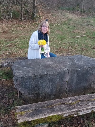 The scene is an outdoor setting within the city of Étalle, Belgium. A blonde-haired female, wearing glasses, a light-colored cardigan, a dark scarf, and blue trousers, is positioned behind a large, dark grey stone block. She appears to be kneeling or seated, with her upper torso and head visible above the block, looking directly at the camera. She holds a bouquet of approximately 15-20 bright yellow flowers, consistent with daffodils, clutched in both hands at chest level. The prominent rectangular stone block in the foreground exhibits a weathered, rough texture with visible cracks and discoloration. Below and in front of the stone block, a moss-covered, weathered wooden plank runs horizontally. The ground around the subjects is covered with patchy green grass and scattered brown leaves. In the background, numerous deciduous trees with bare branches are visible, indicating a late winter or early spring season. The lighting is natural daylight.