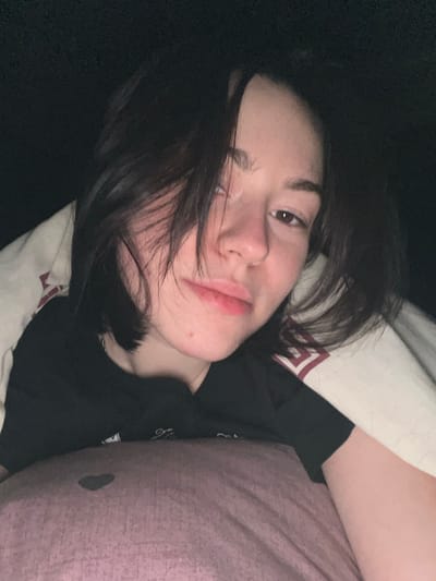 A person with dark hair lies in bed, face tilted towards the camera. Their hair is tousled, partially obscuring their face. The person wears a dark shirt. A white pillow with a red and white pattern is behind their head. A lavender pillow is next to them, slightly stained with a dark mark. The background appears dark, suggesting a dimly lit room.