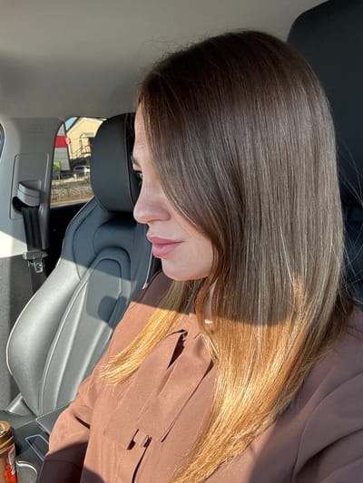 A female individual is depicted in profile, seated in the front passenger seat of a vehicle, looking towards the right. She possesses long, straight, brown hair with lighter, possibly blonde or light brown, ends, strongly illuminated by direct sunlight on her right side. Her skin is fair, and she has light pink lipstick on her lips. She is attired in a brown long-sleeved garment featuring a distinct folded fabric detail on the upper left chest area.

The vehicle's interior includes a black leather passenger seat with white stitching, a black headrest, and a black seatbelt mechanism with a silver buckle visible on the left. An 'AIRBAG' label is visible on the pillar above the seatbelt. A gold-lidded cup or container with a brown label is situated in the lower left of the frame, likely in a cup holder or console.

Through the vehicle's window, an urban outdoor environment consistent with the city of Likino-Dulyovo, Russia, is observable under bright daylight. The scene includes a light-colored building, a vertical red and white striped pole or barrier, a dark metal railing, a paved surface, and a partially visible white automobile.
