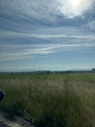 The image captures an expansive rural landscape under a bright, partly cloudy sky. The upper portion of the frame is dominated by a clear blue sky interspersed with numerous thin, horizontal cirrus clouds. The sun is prominently visible in the upper right, emitting a strong glare that diffuses through the high-altitude clouds. Across the mid-ground, a distant, undulating mountain range stretches horizontally, appearing hazy and light blue due to atmospheric perspective. Below the mountains, vast green fields extend across the landscape, with a darker band of trees or dense vegetation visible between the fields and the mountain range. The immediate foreground features tall, dry, light-colored grasses intermixed with greener foliage. A dark, semi-circular object, likely part of a vehicle's mirror or frame, is partially visible on the far left edge. The bottom left also shows a dark strip resembling a road or roadside, with light reflections. The scene depicts an open countryside near Belanitsa, Bulgaria, under bright daylight conditions. No people or animals are visible, and no specific actions or interactions are depicted.