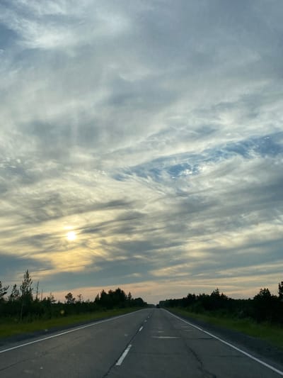 This is a photograph taken from a moving vehicle on a paved road heading towards a horizon line. The road has a white dashed line dividing the lanes and a solid white line on the right edge. On either side of the road are dense green trees forming a border along the horizon. The sky is filled with wispy, streaky clouds in shades of white, gray, and blue, with the sun visible as a bright, diffused orb peeking through the clouds on the left side of the frame. The overall lighting suggests it is either early morning or late evening. The location is the city of Lempino, Russia.
