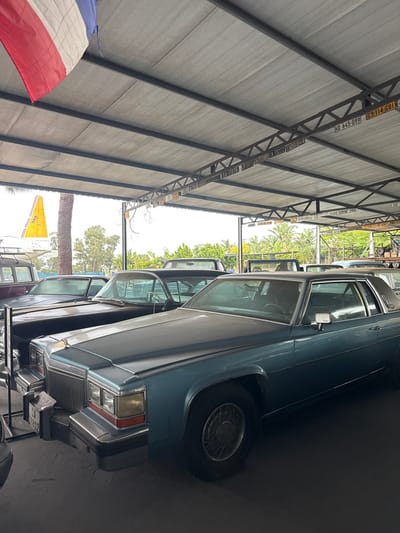 The image shows a row of vintage cars parked inside what appears to be a large garage or storage facility, possibly in Kraaifontein, South Africa, given the license plates. The cars are mostly light-colored sedans, partially covered, with one car taking center stage from a closer perspective. The structure above is made of steel beams supporting a roof. License plates are affixed to the beams. A flag is visible in the left corner. The environment suggests a collection or a storage area for classic cars.