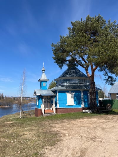 The image shows a blue and white wooden building with a tall pine tree next to it. The building has decorative woodwork and a steeple. The sky is clear and blue. In the background, there is a body of water and some bare trees. A dirt path runs along the right side of the frame. The location is Чернышово, Russia.