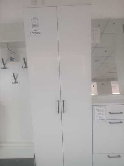The image displays a white wardrobe with two doors. A label is attached to the left door with information about a product, including "Лером", "Прихожая «Грейс»", "Ширина 0,72", "Высота 2,27", "Глубина 0,35", "Россия/ Пенза", and "Цена 170 000". Below the wardrobe, on the left, are several black coat hooks mounted on a white wall. To the right of the wardrobe, a mirrored surface reflects a ceiling with lights and grid patterns. Further to the right, a dresser with three drawers is partially visible. A second label is affixed to the top drawer of the dresser, also mentioning "Лером", "Прихожая «Грейс»", "Ширина 0,54", "Высота 1,19", "Глубина 0,35", and "Цена 130 000". The scene appears to be an interior of a store or showroom. There are no people or animals visible in the image. The city of Jezqazğan, Kazakhstan is not depicted in this image.