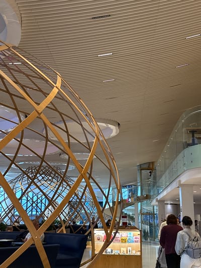 The image depicts the interior of a modern building, likely an airport or a large public space, with a focus on architectural design. In the foreground, a large, spherical wooden lattice structure dominates the view, its curved wooden beams forming an intricate geometric pattern. This structure appears to be a decorative element or part of a larger installation.

Beyond the wooden lattice, the space opens up to reveal a multi-level interior. A sleek, modern ceiling with linear lighting fixtures stretches across the upper part of the image. To the right, a balcony with a glass railing overlooks the lower level, with a spiral staircase and an elevator visible in the distance. People are seen walking and seated in this area, suggesting a functional public space.

In the lower left quadrant, blue upholstered seating is arranged, with several individuals seated and interacting with their surroundings. A display case with various small items is visible near the center of the image. The overall impression is one of contemporary design and spaciousness. The context of the city of Перепечино, Russia, is not discernible from the visual elements present in the image.
