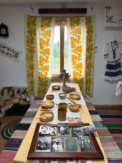 The image depicts an indoor scene, likely a room furnished in a traditional style. The room is decorated with floral curtains featuring orange and white flowers on a light green background, and several pieces of embroidered fabric. On the left wall, a black pendulum clock hangs, and below it, an embroidered runner is displayed. To the right of the window, a white and blue dress with ruffled detailing is hung on a hanger.
In the center of the room, a long table is covered with a light-colored tablecloth. The table is set with various wooden bowls, some containing spoons, and a samovar. There are also several framed photographs on the table, showcasing groups of people in vintage attire, as well as individual portraits.
On the left side of the room, near the window, a fox pelt is laid out on a chest, accompanied by embroidered cloths. The floor is covered with a striped rug. The overall impression is that of a culturally preserved space, possibly a museum exhibit or a well-maintained traditional home. The location is identified as Ust'-Pyzas, Russia, suggesting a cultural context related to that region.