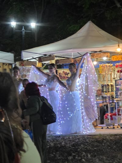At night, two young women in sparkling white dresses are performing a dance. They are holding sheer fabric illuminated by numerous small, bright white lights, creating a magical effect. The women have their hair pulled back and are smiling as they dance. They appear to be on a paved area in front of a market stall. Behind them, stalls are set up, with goods displayed, and a white tent provides overhead cover. The background shows dark trees and streetlights. A person with red hair wearing a green jacket and a backpack is partially visible in the foreground, facing away from the dancers. This scene is located in the city of Gara Oreshets, Bulgaria.