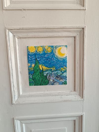The image shows a framed painting, resembling Van Gogh's "The Starry Night," adhered to a white, paneled door. The painting is small and features a swirling blue and yellow sky with a crescent moon, prominent green cypress trees, and a village with a church spire. The door itself is painted white and has a distressed, textured finish with visible brushstrokes and some chipping. The painting is centrally placed within one of the door's panels. The surrounding environment is not visible, only the door and the artwork. There are no people, actions, or interactions depicted. The scene is static and focuses on the decorative element of the painting on the door. The location in Legnica, Poland is not discernible from the visual information provided in the image.