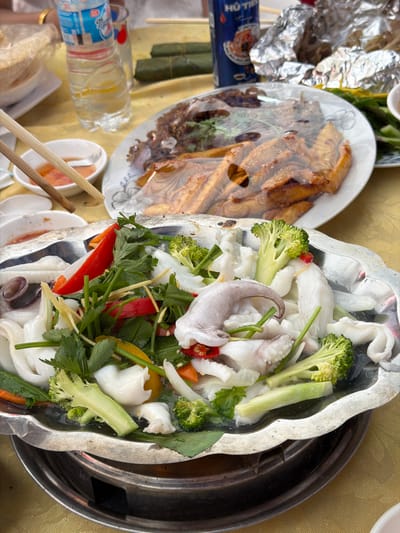 The image captures a vibrant overhead view of a dining table laden with Vietnamese dishes, likely in Tay Son Commune, Vietnam. The central focus is a large, ornate metal platter or pot containing a fresh assortment of ingredients, possibly for a hotpot or steamed dish. This main dish features substantial pieces of white squid, bright green broccoli florets, thin slices of red and yellow bell pepper, slivers of ginger, and various green herbs such as cilantro and parsley. The ingredients appear uncooked or lightly prepared, ready for further cooking at the table.
To the left, a ceramic plate holds several pieces of what appear to be fried or baked goods, possibly fish or snacks, covered with a clear plastic wrap. Adjacent to this, crumpled aluminum foil and some items wrapped in green banana leaves (possibly *banh tet* or a similar snack) are visible.
Beverages on the table include a partially visible blue can with the text "HÔ TÉ" clearly legible, indicating Huda Beer, a popular Vietnamese brand. A plastic water bottle, partially filled, is also present.
The table itself is covered with a golden or light yellow tablecloth. Chopsticks rest on small white dipping bowls, suggesting a communal dining experience. The overall setting is casual, typical of a family meal or a gathering at a local eatery. The bright lighting suggests it is daytime.