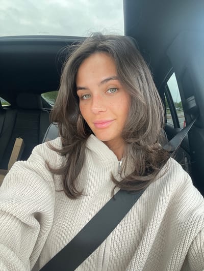 The image shows a young woman with long brown hair sitting in the driver's seat of a car. She is wearing a cream-colored knitted hoodie and a black seatbelt is fastened across her chest. Her right arm is resting on the seat, and her left hand is not visible. She is looking directly at the camera with a soft smile. The background of the image is blurred. The car interior is black. The location of the image is Bradwell Common, United Kingdom, based on the provided coordinates (latitude: 52.04, longitude: -0.776).