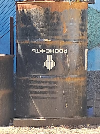 A heavily rusted, black metal barrel stands on a concrete base outdoors. The barrel has the word "РОСНЕФТЬ" (Rosneft) in Cyrillic letters and a stylized white logo resembling a gas flame or an arrow pointing upwards, on its side. The barrel appears weathered and aged, with visible rust streaks and a textured surface. To the left of the barrel, a portion of another, similar barrel is visible. The background includes a textured blue wall on the left, and a textured greenish-gray surface on the right, suggesting an outdoor urban or industrial setting. The ground is covered with a mixture of gravel and debris. The image is taken in Chobruchi, Moldova.