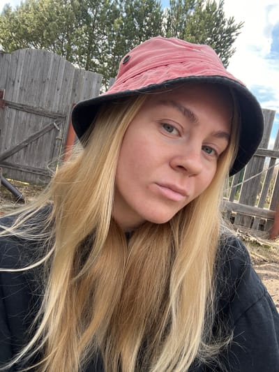 A selfie of a blonde woman wearing a pink corduroy bucket hat with a black brim. She has fair skin, green eyes, and full lips. Her hair is long and straight. She is wearing a black top or jacket. The background includes a weathered wooden fence and a tree. The sky is visible in the upper left corner. The location is likely near Chernysovo, Russia due to the prompt.