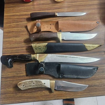 The image displays a collection of six knives arranged in a row on a wooden surface, likely a table. The knives vary in design and appearance, with some having leather sheaths and others displayed without.

From left to right, the first knife is slender with a dark brown handle and a silver blade, partly visible in its brown leather sheath. Next to it is a knife with a brown leather sheath and a silver blade with a brass-colored bolster. The third knife features a black leather sheath adorned with ornate gold metalwork at the tip, and its blade also has decorative etchings. The fourth knife has a black leather sheath with a visible snap closure and a silver blade. The fifth knife has a dark, possibly horn, handle and a silver blade, with its sheath absent. The sixth knife, on the far right, has a light-colored, possibly bone or antler, handle and a silver blade.

The wooden surface has a prominent wood grain pattern. In the background, a sliver of a grey and white tiled floor is visible, along with a glimpse of fabric with stripes of orange, black, and brown, possibly part of clothing or upholstery. The lighting suggests an indoor setting, with no clear indication of time of day or weather conditions. No text is visible in the image. The overall scene is a still-life arrangement of knives.