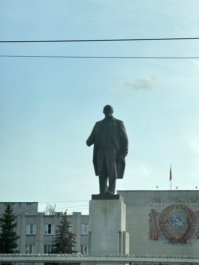 The image depicts a statue of Vladimir Lenin in Kstovo, Russia, standing in front of a building adorned with a Soviet-era emblem. The statue is facing left in the frame and elevated on a concrete platform. A red flag can be seen on top of the building with the emblem. Electrical wires are visible in the sky on the left of the image. An apartment building is adjacent to the statue. The sky is light blue.