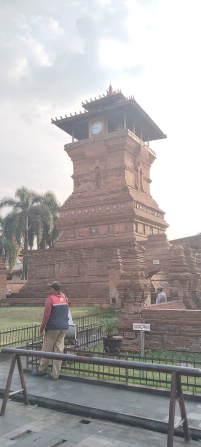 The image shows the Menara Kudus Mosque in Kauman, Indonesia. The mosque is a historic landmark known for its unique architecture, which blends Islamic and Hindu influences. The tower, made of red brick, has a square base and a tiered roof. A clock is visible on one side of the tower, and decorative elements are present on the brickwork. In the foreground, a man wearing a vest and a cap is standing near a metal fence. Palm trees are visible to the left of the tower. The sky is cloudy.