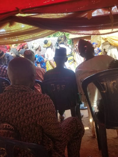 The image portrays an outdoor gathering in the city of Ochadam, Nigeria, observed from within a temporary shelter. A red fabric canopy is suspended overhead, interspersed with draped golden-brown translucent material. The ground beneath is visible as bare earth. In the immediate foreground, three individuals are seated, viewed from behind. The leftmost individual wears a dark, intricately patterned long-sleeved shirt with "TDK" visible on the left shoulder. The central individual is seated on a black plastic chair marked with "JRS" in white on its back, wearing a dark hat and attire. The rightmost individual wears a light-colored traditional garment and a red traditional cap. Beyond these foreground figures, a crowd of approximately twenty individuals is visible, mostly standing and oriented away from the camera. Many attendees are dressed in brightly patterned traditional Nigerian clothing, including head wraps for women and traditional caps for men. Green foliage and a partial view of a light-colored building are discernible in the background, suggesting an open-air setting. The scene is illuminated by bright natural light.