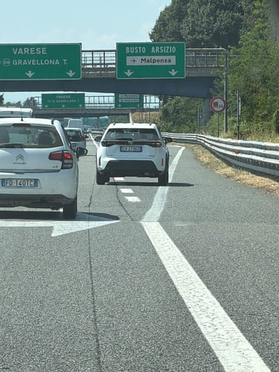 The image captures a view from a vehicle traveling on a highway in Olgiate Olona, Italy, indicated by road signs for "VARESE" and "GRAVELLONA T." and "BUSTO ARSIZIO" with an arrow pointing towards "Malpensa." In the foreground, the rear of a white Citroen C3 with the license plate "FD-140TC" is visible. To its right, the rear of a white Toyota Yaris with the license plate "GV-279RZ" is also prominent. Both vehicles are on a multi-lane paved highway with white lane markings and a metal guardrail along the right side. In the background, overhead road signs and lush green trees line the highway, suggesting a sunny daytime environment. A circular traffic sign depicting a truck within a red circle and a diagonal line is visible on the right side of the road. The overall scene depicts normal highway traffic flow under clear weather conditions.