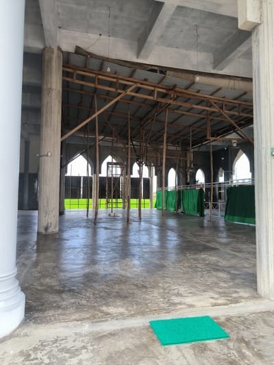 The photo shows the interior of a large, open structure, possibly a mosque, in Blangbitra, Indonesia. The building features a concrete floor and a partially exposed ceiling with wooden supports. Arched windows are visible along the back wall, offering a view of a green field. Green curtains and scaffolding suggest ongoing construction or renovation. Several pillars support the roof, and a green mat lies on the floor near the entrance. No people are visible in the image.