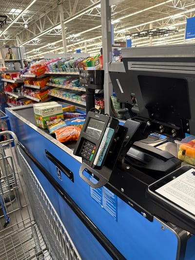 The image, correctly oriented, displays an interior view of a retail store, specifically a checkout lane at what appears to be a Walmart. The brightly lit environment features a high, exposed ceiling with white beams and fluorescent lighting.

In the foreground and midground, a blue checkout counter is visible. Attached to the counter is a payment terminal, branded "Walmart" and "Ingenico," with a numerical keypad and a screen. A blue "Walmart" gift card or similar payment card is partially inserted into the terminal's chip reader slot, suggesting a transaction is either in progress or about to occur. Next to the terminal, a clear plastic container holds what appears to be pre-cut vegetables, possibly carrots, indicating a purchase. Above this, a tablet or screen displays illegible text, likely instructions or transaction details. A scanner or scale mechanism is also integrated into the counter setup. A metal shopping cart is partially visible on the right, positioned as if waiting to be loaded.

Behind the checkout area, several shelves are stocked with various packaged snack items, including chips, candies (like Reese's), and cookies (e.g., Goldfish crackers, Chex Mix), often placed as impulse buys. A price tag displays "7.49". Further in the background, more store aisles with merchandise are visible, extending into the distance. A blue sign in the upper left background is partially visible, with "Self Checkout" being faintly decipherable, confirming the type of service area. Another rotated sign in the far background displays the "Reebok" logo.

No people are visible in the image. The scene captures a moment at an active retail checkout station, focusing on the equipment and merchandise. The prominent "Walmart" branding on the payment terminal and card, along with the "Self Checkout" sign, clearly identify the location as a Walmart store in the United States. The indoor setting provides no information regarding the time of day or external weather conditions.