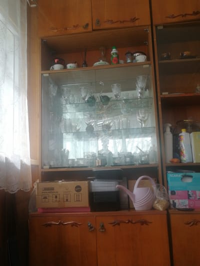 A close-up, eye-level shot captures a cluttered wooden cabinet in what appears to be a domestic setting. The cabinet is filled with an assortment of glassware, bottles, and boxes. On the top shelf, a green bottle with a red rose label is visible, alongside other small containers and a ceramic cup. The middle section features numerous clear glass items, including wine glasses, tumblers, and decorative pieces, arranged on glass shelves. Below, a cardboard box with Russian text and shipping symbols sits on a pink surface. To the right of this box, a light pink watering can and a plastic bag filled with what appears to be grain are placed on a shelf. Further to the right, a blue box with "SCARLETT" branding is visible. The overall impression is one of storage and organization of various household items within a wooden cabinet. The scene is likely located in Dubrovka, Russia.