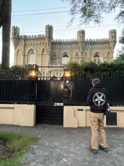 A vertically oriented photograph captures a scene at what appears to be dusk or early morning, given the soft, diffused light and an illuminated street lamp. The image centers on a person standing on a textured paved surface beside an ornate black metal gate, with a distinctive castle-like building dominating the background.
The individual, seen from behind, is wearing a black hooded sweatshirt and light-brown (khaki) pants. A prominent white circular emblem is emblazoned on the back of the hoodie. This emblem features a five-pointed star (pentagram) that contains a stylized goat's head, widely recognized as the Sigil of Baphomet or related occult symbolism. The person has short, dark hair and is wearing dark shoes.
Behind the intricate black metal gate stands a large, historic-looking building with a fortified or castellated architectural style, characterized by multiple crenellated towers, arched windows, and detailed stone work on its facade. Some windows are fitted with white shutters. Green foliage, including trees and bushes, is visible around the building and partially frames the sky. Utility lines stretch across the upper portion of the sky. The ground consists of dark, uneven paving stones, with a small patch of green grass growing alongside a light-colored wall segment.
The setting, identified as Olivos, Argentina, presents an imposing yet somewhat mysterious ambiance due to the historical architecture and the distinct symbol on the person's attire. No other discernible text is visible in the image.