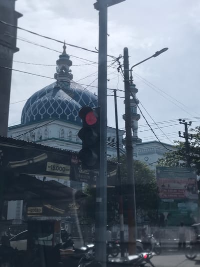 A daytime street scene captures a mosque with a distinctive blue patterned dome and minarets, viewed from what appears to be inside a vehicle, as evidenced by significant reflections across the image. The sky is overcast, and the scene is partially obscured by street infrastructure including lamp posts, utility poles with numerous wires, and a traffic light displaying a red signal.

The mosque features light-colored walls and arches below its prominent blue dome, which is adorned with an intricate, wave-like pattern. Several ornate minarets, topped with pointed finials, rise alongside the dome. In the foreground, a dark-colored traffic light hangs with its red signal illuminated.

Visible reflections on the car window include trees, parts of other buildings, and a large billboard or banner. Text on this reflected banner, when deciphered and rotated, advertises "HEWAN QURBAN" (Sacrificial Animals), indicating an offering for Eid al-Adha. Further details state "PANITIA QURBAN" (Qurban Committee), "Menerima Hewan Qurban" (Receiving sacrificial animals), and pricing: "1 Rp. 2.500 000 / Orang" (Rp 2.5 million per person) and "2 Rp. 3.000 000 / Domba" (Rp 3 million per sheep). Other reflected text mentions "MAGNUM KRETEK" (likely a cigarette brand), "BARU" (new), and "RP15.000-an" (around Rp 15,000). The presence of Indonesian Rupiah and the "Qurban" theme strongly suggest a setting in Indonesia, specifically Kutalhoksukon as per the provided context. No people are clearly visible, but the urban setting implies activity.