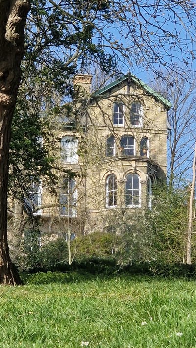 A three-story brick building is partially obscured by trees. The building is light brown in color and has a green roof. It features rectangular and arched windows. The surrounding environment includes green grass and trees with bare branches. The image was likely taken in the city of Lincoln, United Kingdom.