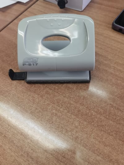 A light blue hole punch with the model number "P-817" visible is positioned on a wooden desk. The hole punch is centrally located in the frame, with its open slot visible. A small black plastic piece, possibly a guide or debris catcher, is attached to the side of the hole punch. The desk surface has a wood grain pattern with variations in color and texture. In the upper right corner of the image, a portion of a smartphone with a purple case is visible, and in the upper left, a white object and a black object are partially visible. The scene is consistent with an office or workspace environment, and there is no information available to determine its location in Dobrodan, Bulgaria.