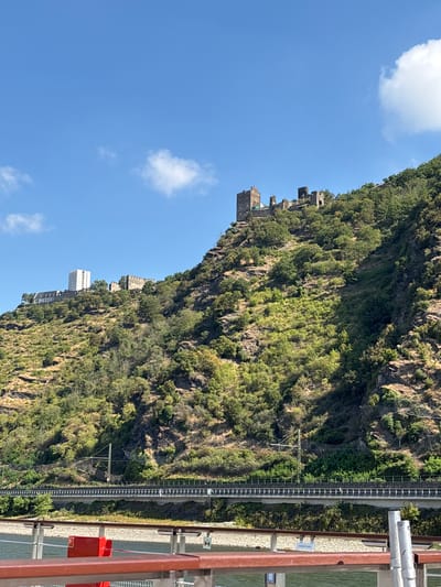 The image shows a hillside in Boppard, Germany, with a river in the foreground and a castle ruin at the top. The hillside is covered in trees and vegetation, and there is a train track running along the side of the hill. A modern building with scaffolding is visible on the left side of the hill, next to an older building. The castle ruins are weathered and appear to be made of stone. The sky is blue with a few scattered clouds. The river is wide and calm, with a gravelly bank. In the foreground, a railing and table are visible, suggesting the photo was taken from a boat or a riverside establishment.