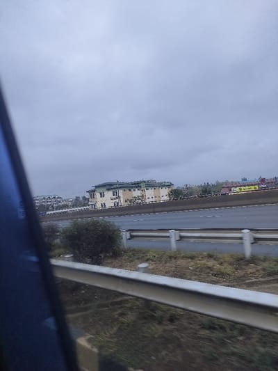A view from a moving vehicle shows a highway with guardrails. Across the highway is a large building with a light-colored facade and green roof, likely an office complex. To the left, a few apartment buildings are visible. A small bush is positioned between the guardrails and the highway. The sky is overcast. The foreground shows the interior of the vehicle, with a blue window frame visible.