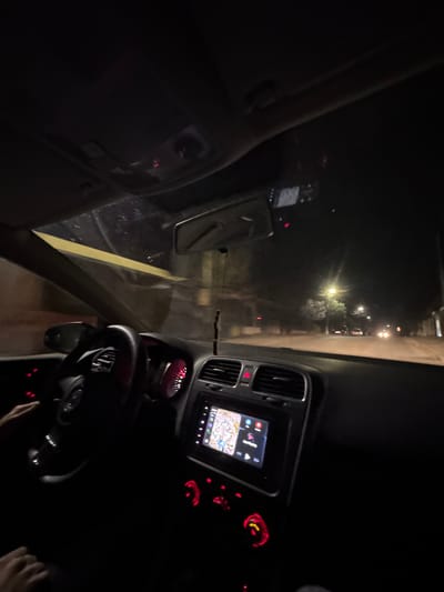 The image depicts the interior of a vehicle at night, viewed from the passenger side, looking forward through the windshield while in motion on an urban street in Kumanovo, North Macedonia. The driver's left hand is visible on the black steering wheel at approximately the 9 o'clock position. The vehicle's dashboard is illuminated with red light, highlighting the instrument cluster and various controls. A central infotainment screen displays a colorful navigation map. Below this screen, a series of red-lit rotary dials and push-buttons are present. A rearview mirror is affixed to the dark overhead console, which contains faint red indicator lights. A horizontal strip of yellow-orange material is applied to the upper portion of the windshield. Outside the vehicle, the environment is dark, with an urban street illuminated by multiple yellow-orange streetlights. Silhouettes of trees are visible along the left side of the road, and the headlights and taillights of several distant vehicles indicate active traffic.
