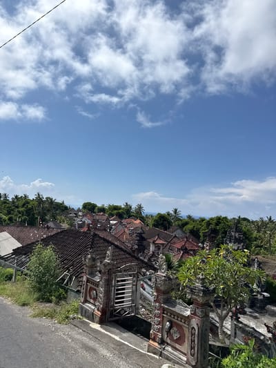The image presents a view in Pejukutan, Indonesia, showing a blue sky with scattered clouds taking up the left half of the frame. The right half consists of traditional Balinese architecture, including residential buildings with reddish-brown tile roofs intermixed with lush green trees and vegetation. A weathered concrete wall and gray asphalt road are visible in the foreground. The structures have ornate detailing and decorative elements.