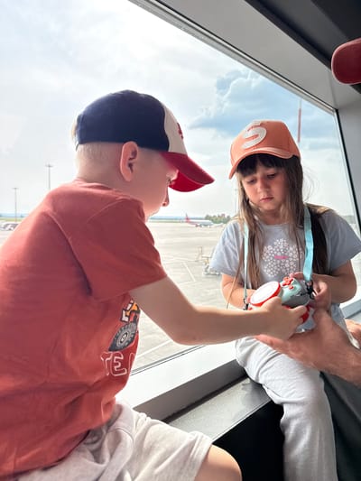 At an airport in Bolshiye Kabany, Russia, two children sit by a large window. The child on the left, wearing a red shirt and a baseball cap, leans in to look at a toy camera held by the child on the right. The child on the right, wearing a light blue t-shirt and an orange baseball cap, gazes down at the camera. An adult's hands are visible, assisting with the camera. Outside the window, airplanes and airport infrastructure are visible under a cloudy sky.