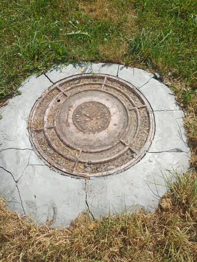 This media file captures a close-up, overhead view of a circular, cast-iron manhole cover set into a cracked concrete surround. The manhole cover is weathered and shows signs of rust, with a circular, raised pattern and a smaller circular emblem in the center. Some dry grass and dirt are lodged within the grooves of the manhole cover. The concrete surrounding the manhole is broken and shows several significant cracks, with patches of green grass growing on one side and dry, brown grass on the other. The scene appears to be outdoors, likely in a grassy area. The lighting suggests it is daytime, with shadows cast by the grass, indicating a sunny day. The location context provided is Elitnyi, Russia, which aligns with the visual cues of outdoor vegetation. No people are visible in the image. The emblem on the manhole cover has some visible text, possibly related to a standard or manufacturer, appearing to be in Cyrillic.