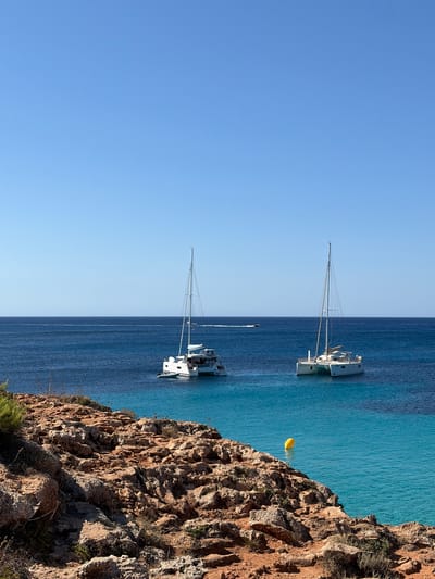 The image depicts a bright, sunny day with a clear blue sky above a tranquil body of water. In the foreground, a rocky, reddish-brown coastline is visible, sparsely covered with dry vegetation. The water transitions from a vibrant turquoise near the shore to a deep blue further out.

Anchored in the blue water are two white catamarans, each with two hulls and tall masts. The catamaran on the left appears to have a smaller dinghy attached. Both boats are stationary.

In the middle ground, a yellow buoy floats on the surface of the turquoise water, indicating a marker or mooring. In the far distance, a faint white line suggests a distant boat or wake on the horizon. The overall scene conveys a sense of peacefulness and natural beauty. This scene could be located along the coast of Sant Lluís, Spain, a municipality on the island of Menorca known for its picturesque coves and clear waters.