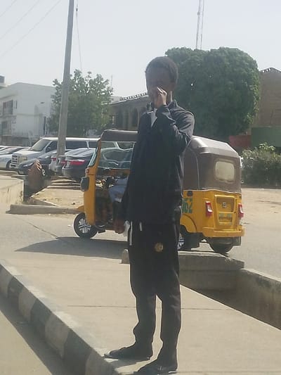The scene depicts an outdoor urban environment in Gyadi Gyadi Arewa, Nigeria, under clear daylight conditions. A dark-skinned individual, appearing male, is standing in the foreground on a concrete sidewalk. This individual is dressed in a black long-sleeved top and black track pants featuring a white stripe on the left leg and a circular emblem on the upper thigh. Their right hand is raised to their face, obscuring the mouth and lower nose area, while their left hand holds a small white object. The sidewalk has a raised curb with black and white striped markings.
In the mid-ground, a yellow and brown three-wheeled auto-rickshaw, commonly known as a "Keke Napep," is visible on a paved road, facing away from the camera. A number "162" is painted on its rear. A person is partially visible inside the driver's compartment. Behind the auto-rickshaw, several vehicles are parked, including a white SUV, a dark sedan, and a white stretch limousine-like vehicle, positioned beyond a low concrete barrier.
The background features a multi-story white building on the far left and an older building with arched architectural details. Sparse green trees and shrubs are scattered throughout the background, with a prominent large, round-canopied tree on the right side. A tall, grey utility pole with overhead wires is visible on the left, extending towards a bright, somewhat hazy sky. Shadows are cast by the subjects and structures, indicating direct sunlight.