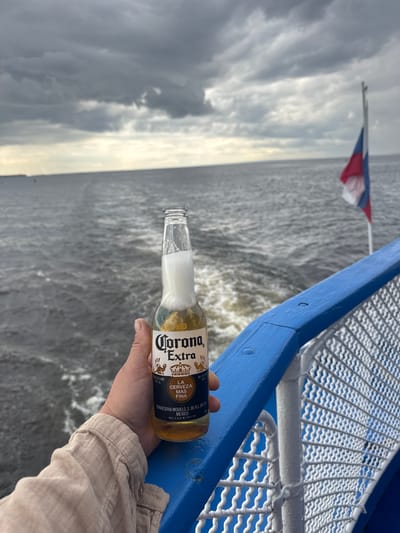 A person's hand holds a bottle of Corona Extra beer with foam spilling out of the top. The bottle is positioned in the foreground, against a backdrop of a grey, cloudy sky over a dark, choppy body of water. A blue railing with a white mesh fence runs along the right side of the image. In the distance, a flag with red, white, and blue stripes flies from a pole. The scene appears to be on a boat or ferry. The location is identified as the city of Belous, Russia.