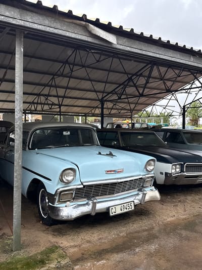 The image shows a light blue classic car parked under a metal-roofed structure in Kraaifontein, South Africa. The car has a "CJ 49455" license plate. Another, darker car is partially visible behind it. The ground is wet and muddy. The metal structure is a simple, functional design with exposed beams. The weather appears overcast.