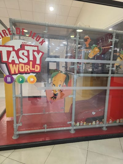 A glass-enclosed display case featuring cartoon characters hangs in an indoor space. "Aire De Jeux Tasty World" is printed on the glass with a logo that includes orange, green, and purple circles. The case's frame is made of grey pipes. A red background is partially visible through the glass. The surrounding area consists of tile flooring and walls, suggesting a commercial environment such as a mall or restaurant.