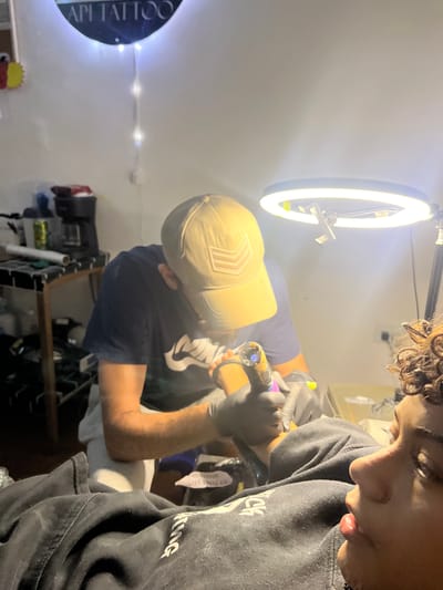 This image depicts a tattoo artist working on a client's arm within what appears to be a tattoo studio. The artist, wearing a beige baseball cap and a navy blue t-shirt, is focused on the client's forearm, holding a tattoo gun. The client, a young person with curly hair, is lying down and looking upwards. The environment includes a bright ring light illuminating the workspace, shelves with various items in the background, and a sign that reads "API TATTOO" on the wall. The overall impression is one of a professional tattooing session. There is no information in the image to place this scene in San Antonio, Venezuela.