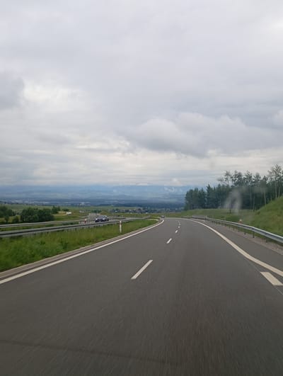This image shows a view from a moving vehicle on a highway that curves to the left, passing by a grassy embankment with trees and a distant valley with a town and mountains under a cloudy sky. The highway is paved with asphalt and marked with white lines. A silver car is visible on the highway in the distance, and a few other vehicles are also present. The sky is overcast with grey clouds. The location is identified as being in Klikuszowa, Poland.