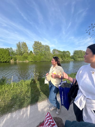 The image depicts two people standing on a path next to a canal or waterway in what appears to be a park. The location is Marquette-lez-Lille, France.
One person, wearing a white top and dark bottoms, is partially visible at the top of the frame. The second person, below them, is wearing a light-colored t-shirt and blue jeans, and holds a blue shopping bag with the name "grandfrais.com" printed on it. They also carry a beige handbag. Both individuals are looking downwards, seemingly at the water.
The waterway is lined with green vegetation, with trees along the far bank. The sky is blue with wispy clouds. Part of a red and white striped object is visible on the right side of the path.