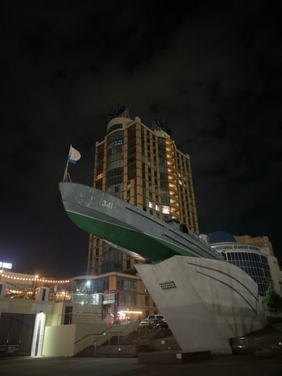 This nighttime image captured in Varnavinskoye, Russia, features a striking monument of a submarine, likely a memorial or landmark, prominently displayed in front of a modern, illuminated high-rise building. The submarine, painted gray with a green hull, is mounted on a concrete base that is designed to resemble the bow of a ship. A flag, predominantly white with blue and red elements and a circular emblem, flies from the submarine's periscope. The number "341" is visible on the side of the submarine.

The tall building behind the submarine has a multi-story facade with numerous windows, some of which are lit, suggesting occupancy. The building's architecture is modern, with a curved top section and several antenna-like structures. To the right of the main building, another structure with a blue dome is partially visible. In the foreground, a paved area with stairs and railings suggests a public access point. Scattered lighting fixtures, including strings of lights on a lower building to the left, illuminate the scene. The sky is dark and appears to be cloudy, with faint hints of stars or distant lights. No people are clearly visible in the image, and there are a few parked vehicles in the background. The overall scene suggests a public space with a focus on maritime history or commemoration.