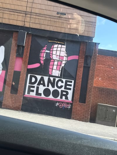 The image displays the exterior of a building in Preston, United Kingdom, captured from an elevated perspective, likely inside a vehicle, evidenced by the lower frame and reflections. The primary subject is a large mural applied to a black surface on the building's facade, which is primarily constructed of red-brown brick.
The mural's upper section features abstract, stylized artwork composed of pink and white checkered spherical shapes connected by a curving pink line. This artwork exhibits some signs of wear, including minor tearing or peeling. Directly beneath this, in prominent white block capital letters, is the word "DANCE" stacked above "FLOOR," with a thin pink horizontal line underneath the text. At the bottom right of the black mural section, a graffiti tag is visible, rendering "#REASM" in white with blue detailing.
To the right of the mural, the building wall continues with exposed red-brown brickwork. A grey utility box, approximately waist-height, is affixed to this brick section, bearing the number "56." Above the main facade, an upper section of the building features a uniform pattern of light beige rectangular tiles or bricks. A thin dark wire descends vertically from this upper section.
The ground directly in front of the building consists of light grey pavement. A clear blue sky is visible in the upper right quadrant of the image, although parts of the upper building and sky are partially obscured by reflections on the glass through which the photograph was taken. No individuals are visible in the scene. The lighting suggests a bright, sunny day.
