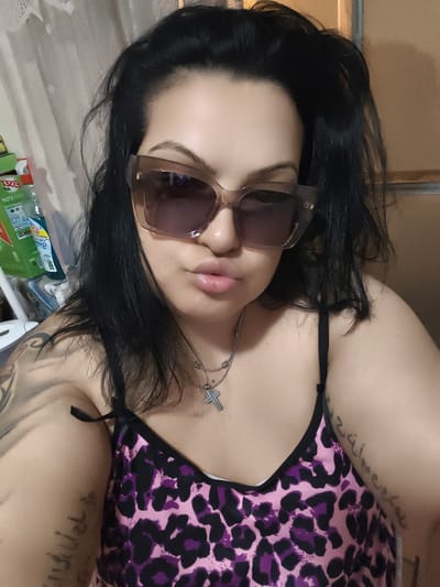 A woman with dark hair and sunglasses is taking a selfie in what appears to be a domestic setting, possibly her home. She is wearing a camisole with a pink and black leopard print. A silver necklace with a cross pendant is visible. There are tattoos on her arms and shoulder. In the background, there are some cleaning supplies and a curtain. The lighting is artificial, creating some shadows and highlights. The image does not provide any information to place it in Komotini, Greece.