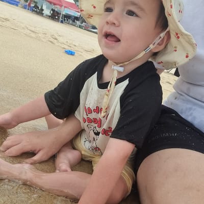 A close-up, slightly tilted shot captures a young child sitting on a sandy beach in Gangneung-si, South Korea. The child, wearing a light-colored sun hat with a cherry pattern and a black and white raglan t-shirt, is looking up and to the right with their mouth slightly open and eyes wide, seemingly engaged with something out of frame. Water droplets are visible on the child's face and clothing, indicating they have been playing in or near the water. Their legs, also wet, rest on the sand.
In the background, out of focus, is the beach environment. Hints of structures like tents or canopies can be seen, along with what appear to be beach chairs or stalls, suggesting a populated beach area. A small, bright blue plastic shovel lies on the sand to the left of the child. The lighting suggests it is daytime, possibly mid-morning or afternoon, with bright, natural light. The overall mood is one of casual enjoyment of a beach day.
On the child's t-shirt, a graphic is visible which appears to be a character, possibly Mickey Mouse, with red text that is partially obscured and difficult to read clearly. Some of the text may say "FOOD". There is also some unreadable text in white on the child's black shorts.